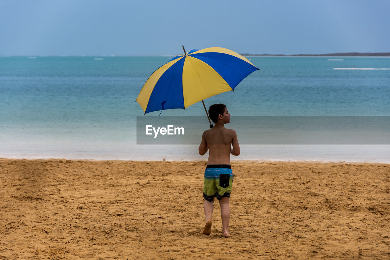 Rear view of boy walking on beach with umbrella