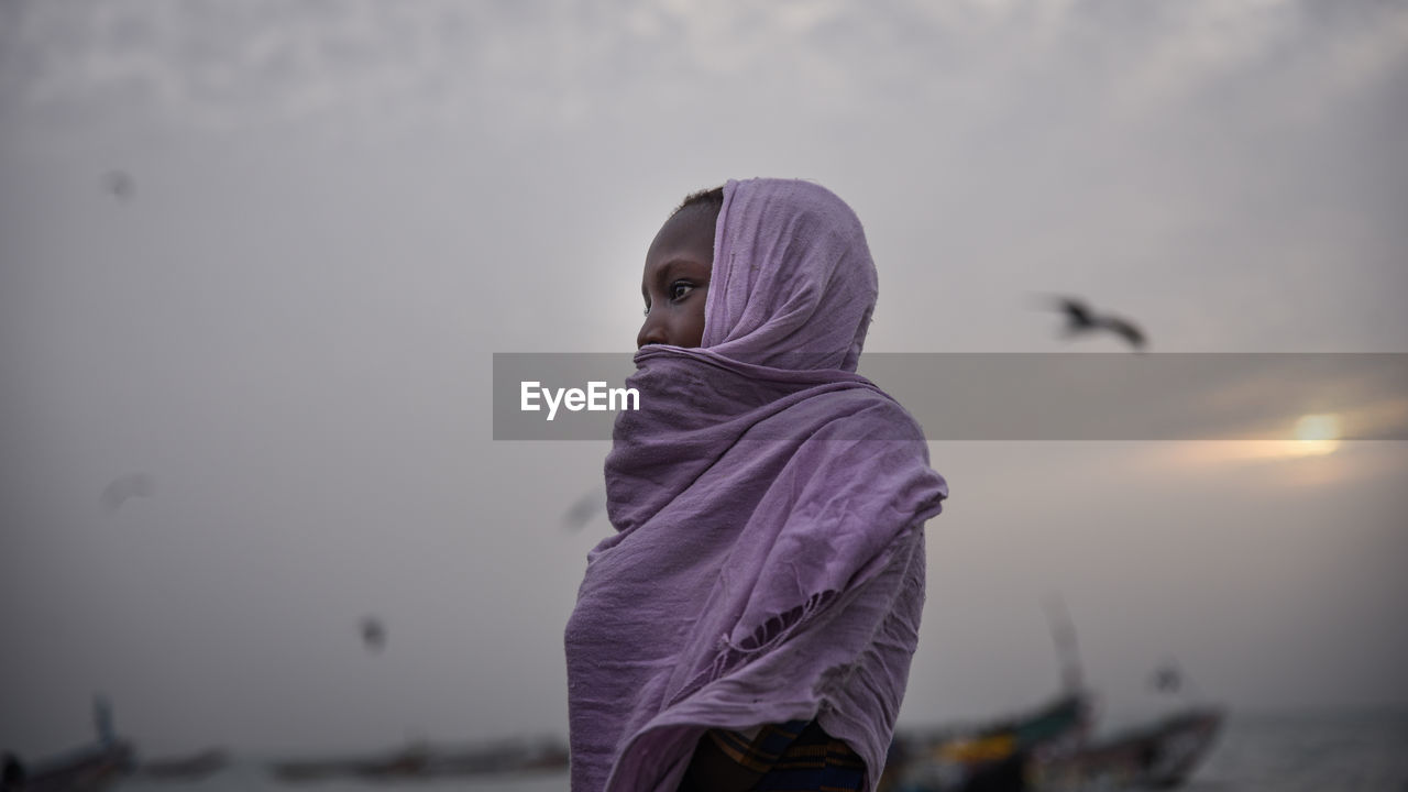 Side view of woman wearing scarf standing at beach against sky during sunset