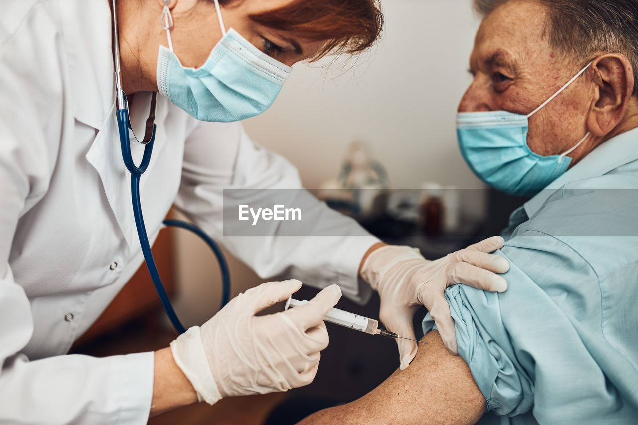 Doctor holding syringe with vaccine and making injection to senior patient with medical mask