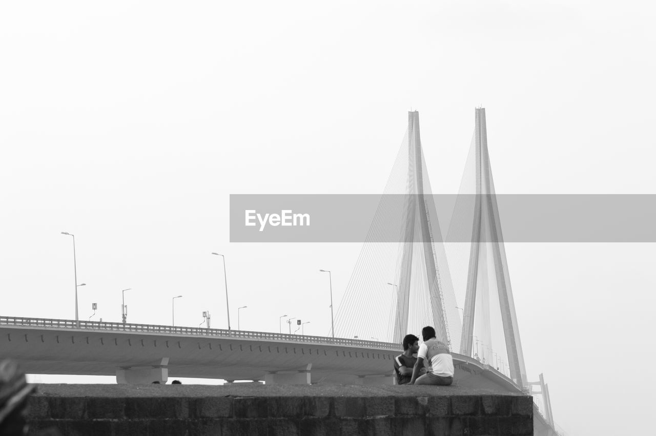 LOW ANGLE VIEW OF MEN STANDING ON BUILT STRUCTURE AGAINST CLEAR SKY