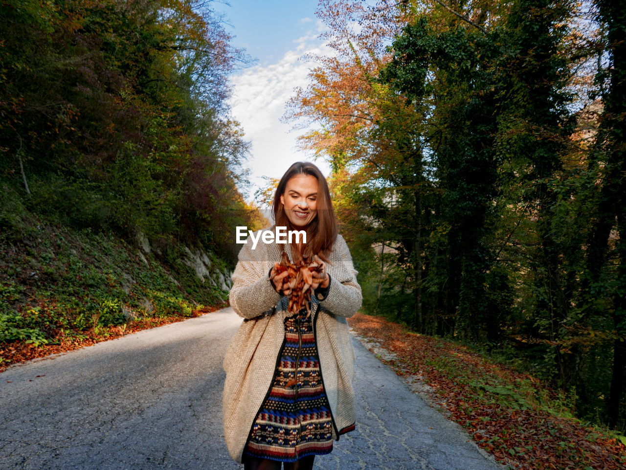 Smiling woman holding leaves while standing on road against trees