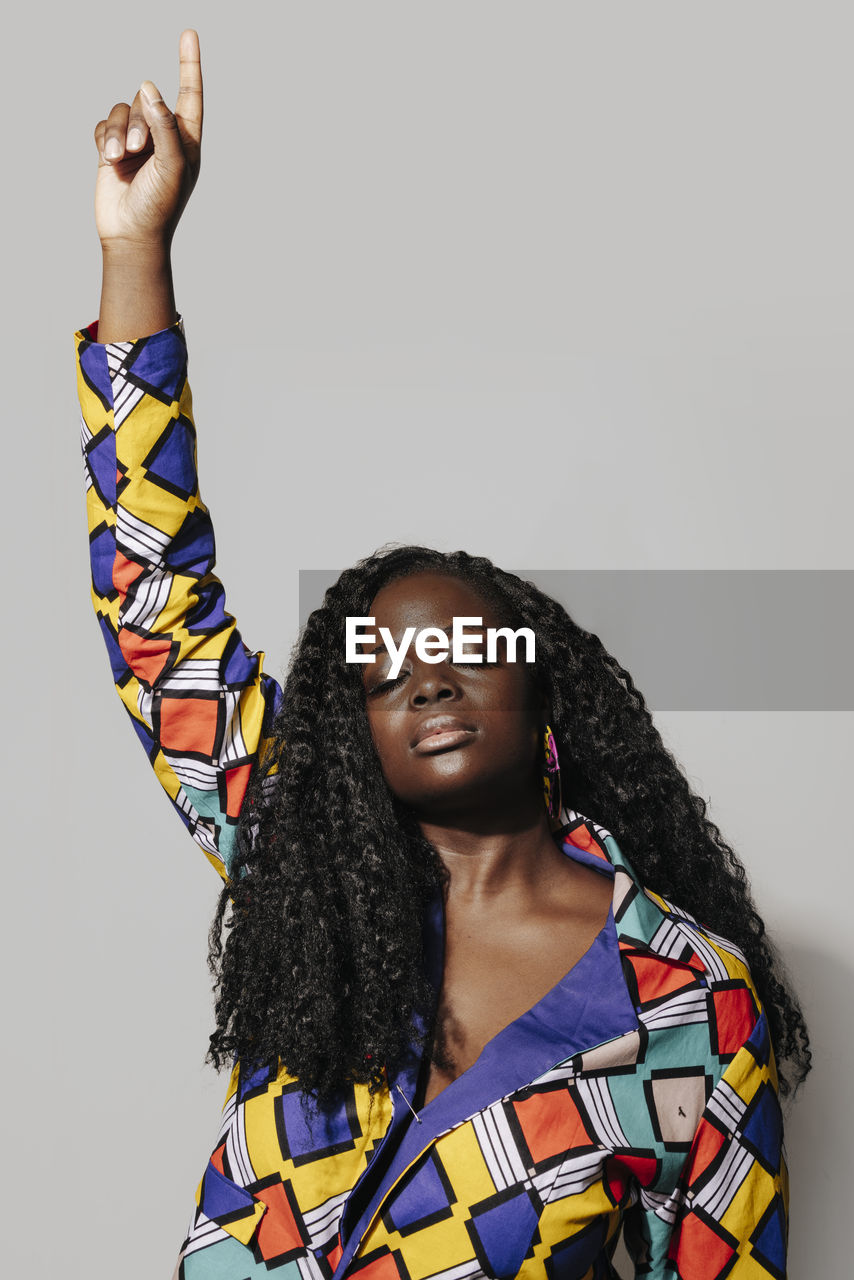 Pensive african american woman with curly hair in stylish african outfit standing on white background. she is with eyes closed pointing finger up