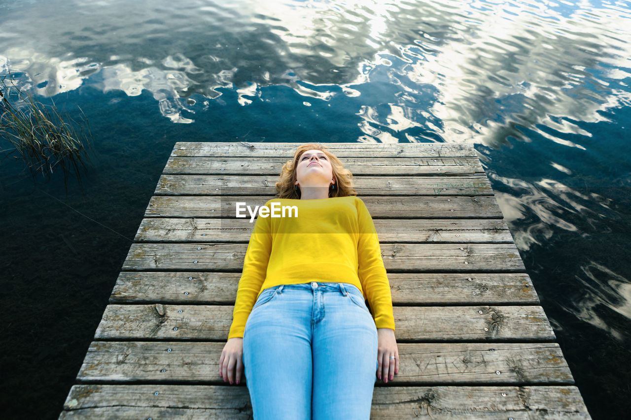 High angle of tranquil female in casual wear lying on wooden pier near lake and enjoying silence on summer day