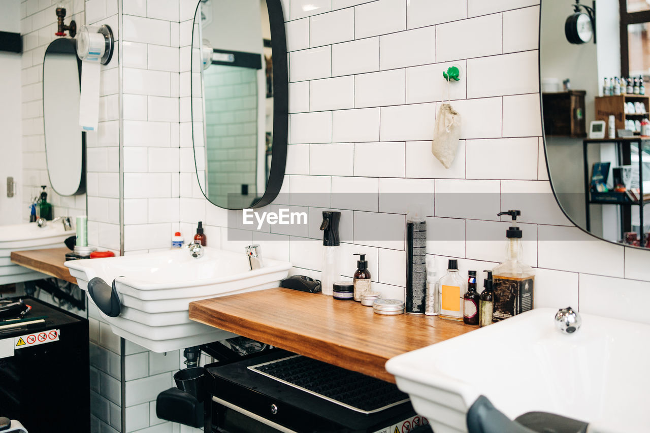 Table with assorted cosmetic products in bottles and dispensers between washstands under mirrors reflecting barbershop