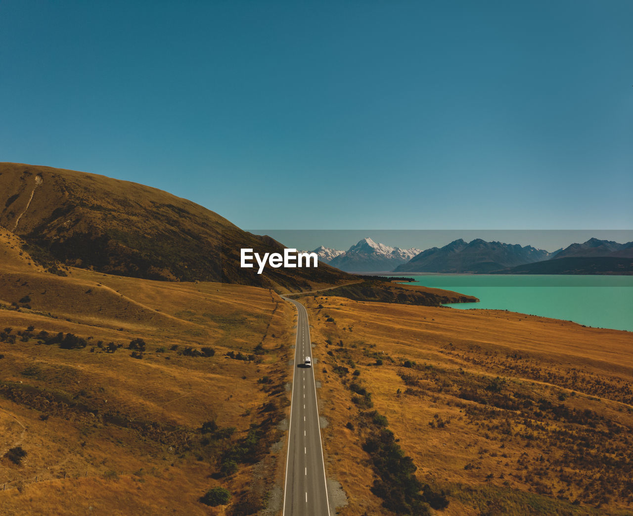 Scenic view of road by mountains and lake against clear blue sky