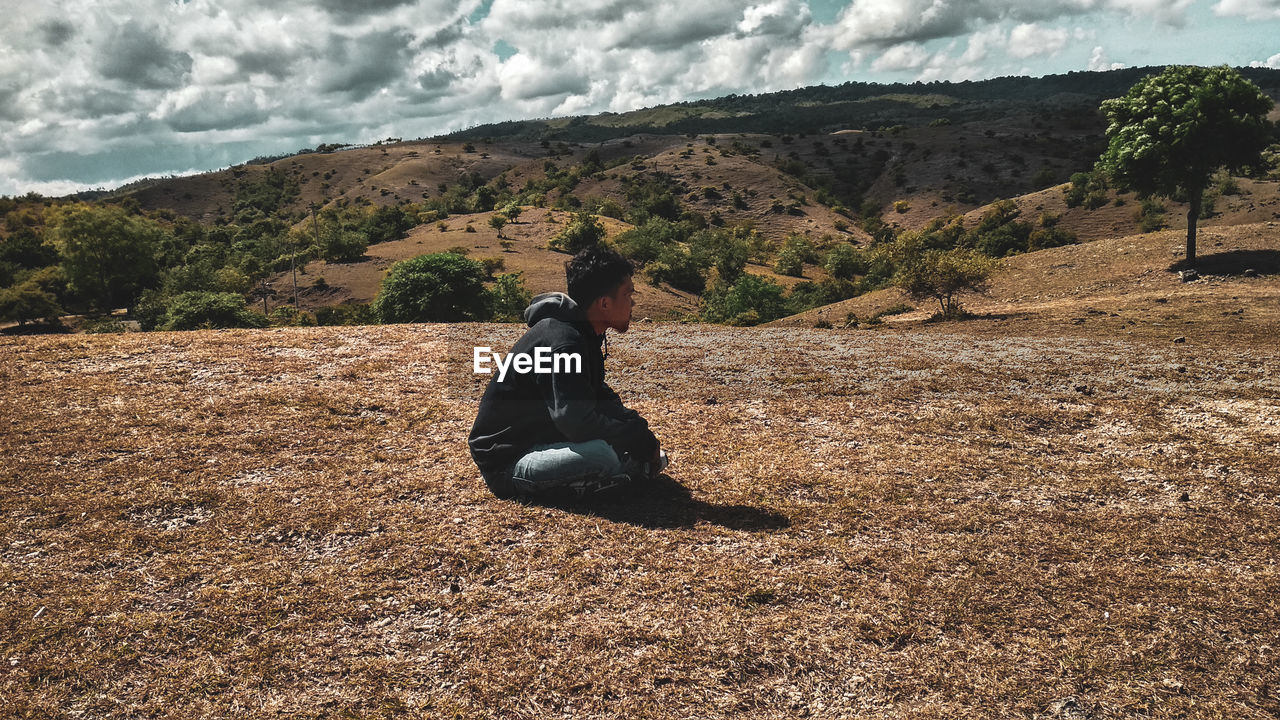 Side view of man sitting on landscape against cloudy sky