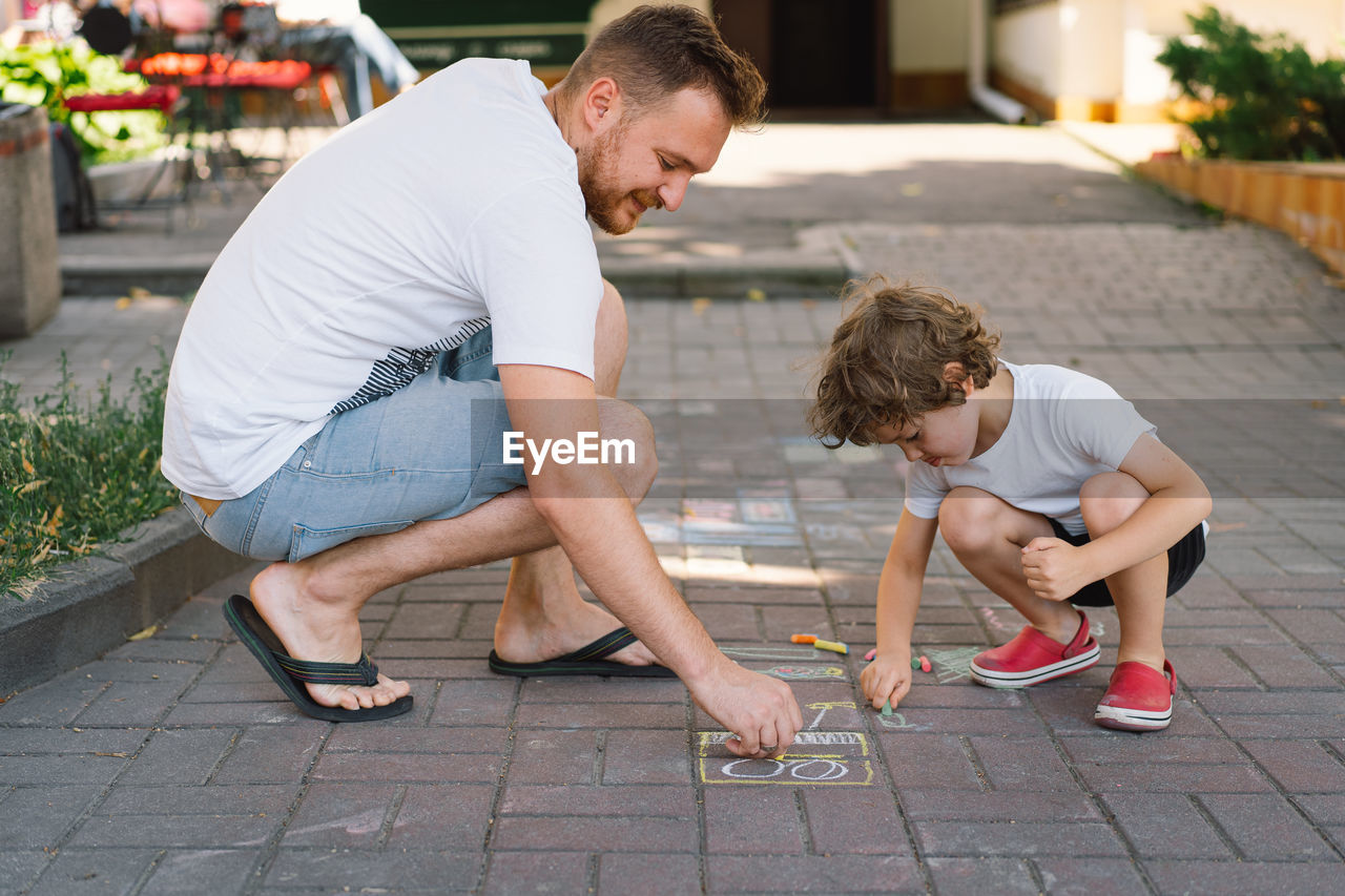 A little preschool boy with father draws with colorful chalks on the ground.