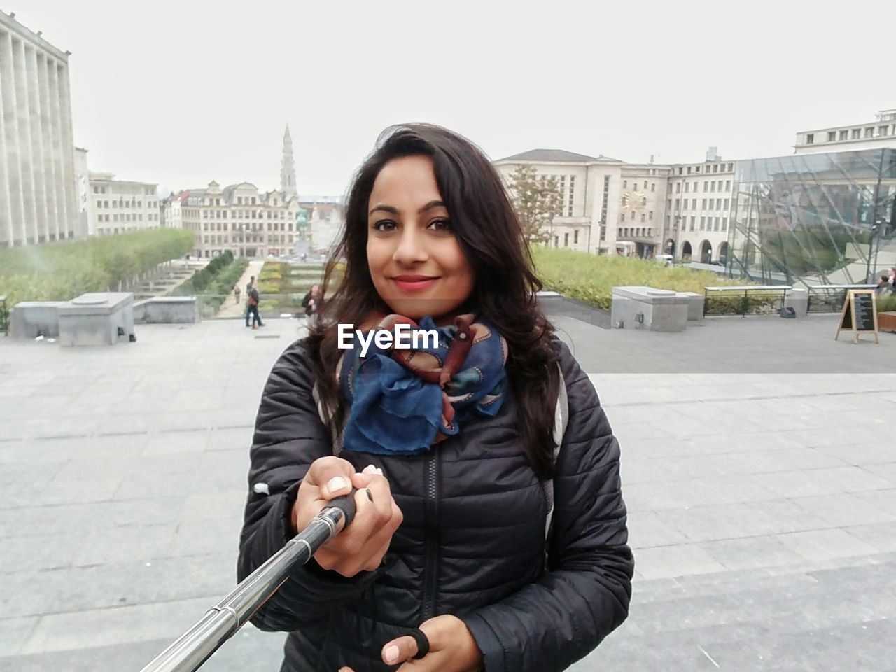 Portrait of woman holding monopod while standing against buildings in city