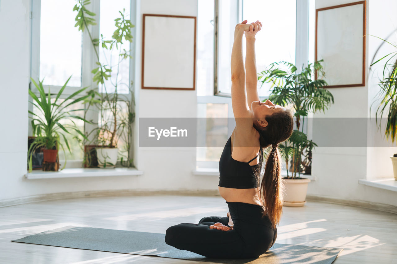 Young fit woman practice yoga doing asana in light yoga studio with green house plant