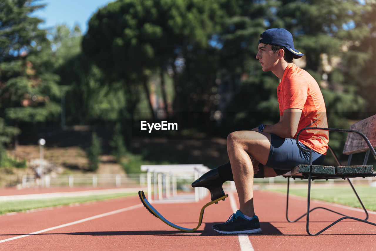 Side view of young athlete with prosthetic leg sitting on bench at running track