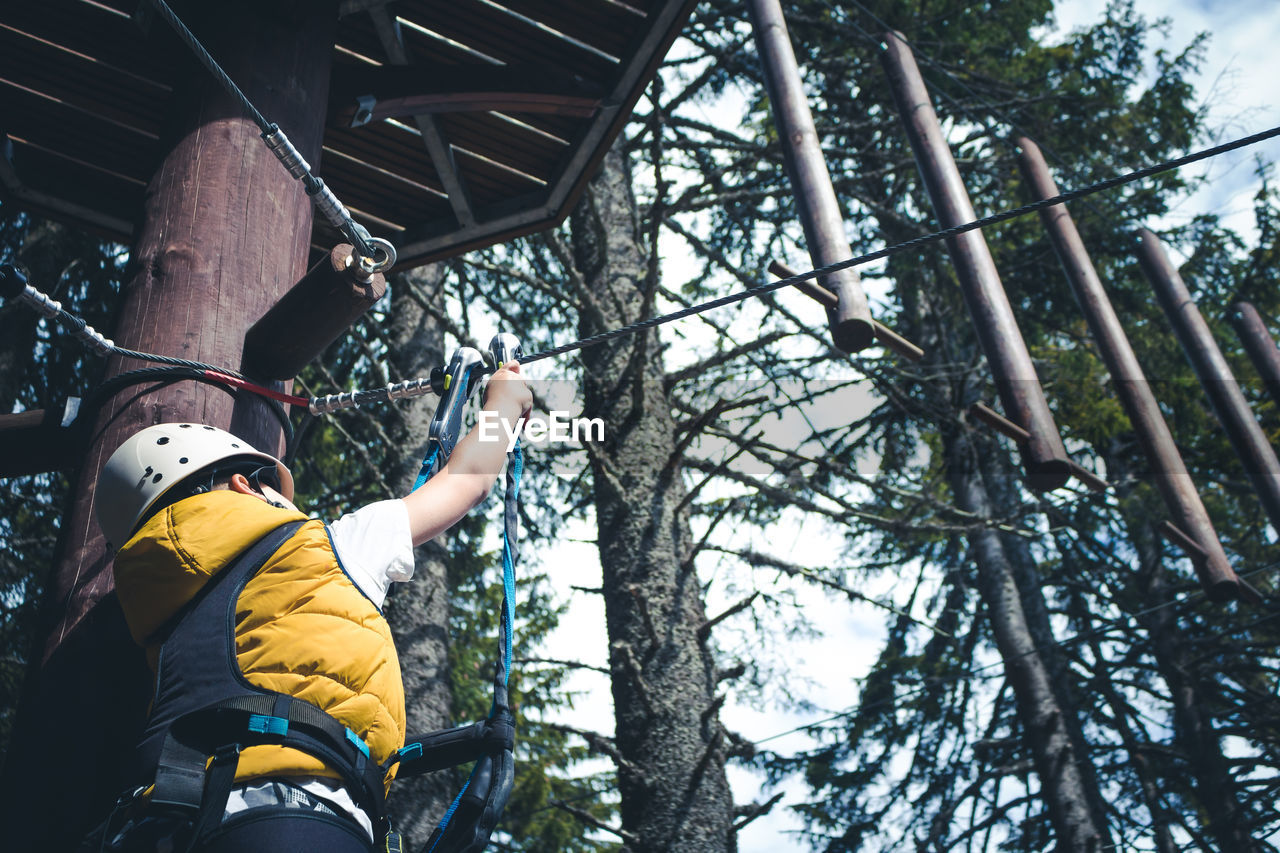 Below view of kid attaching safety harness on zip line while standing on treetop in adventure park.