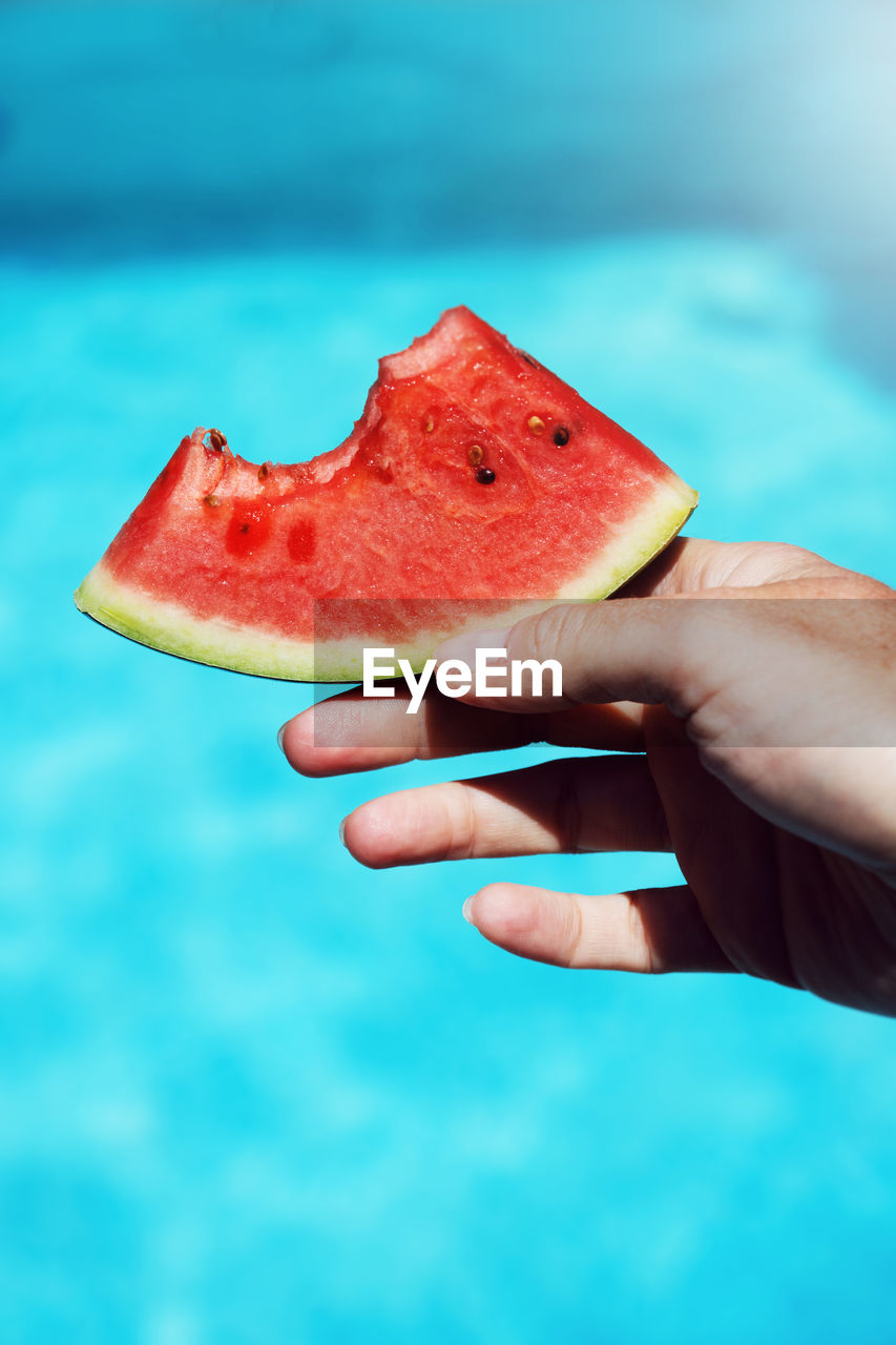 Cropped hand holding melon slice against swimming pool