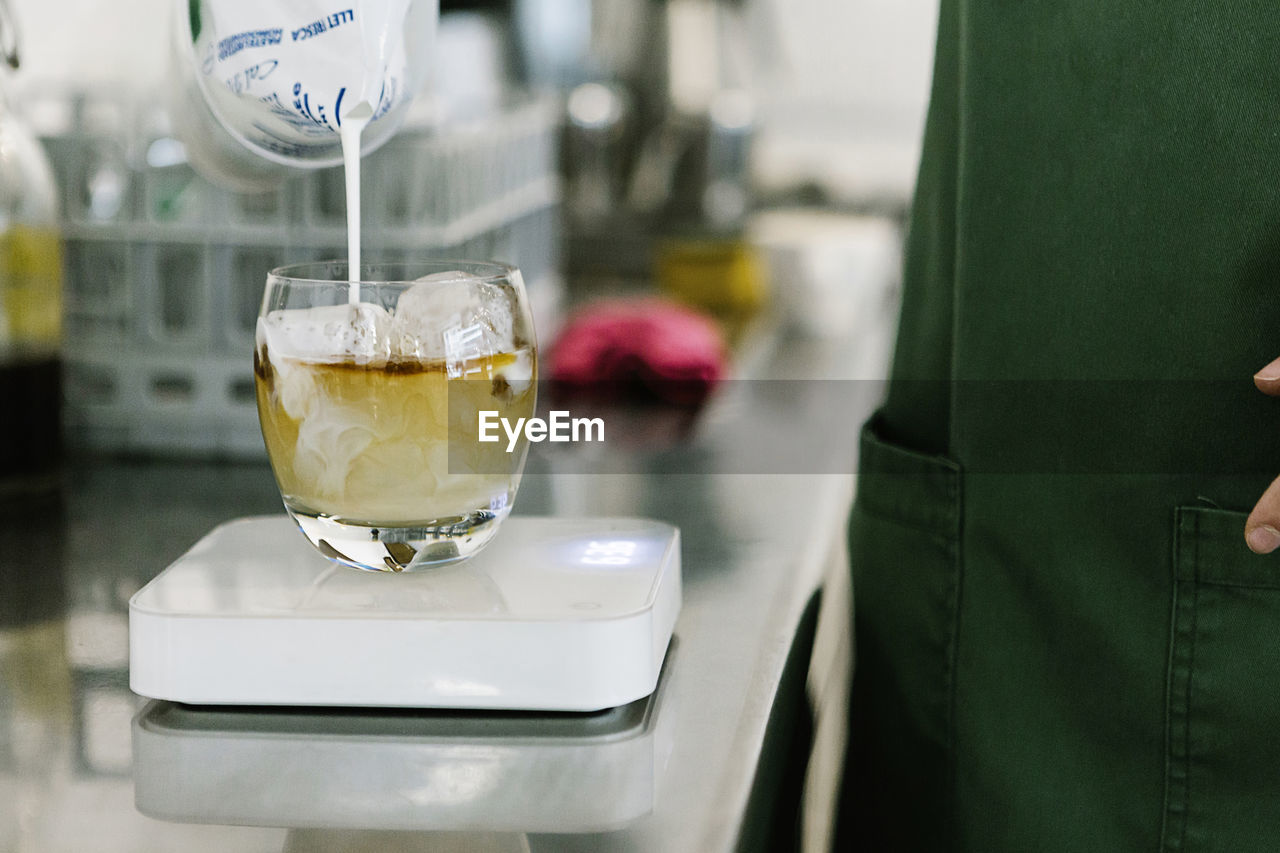 Barista standing while pouring milk in coffee cup kept on kitchen scale at coffee shop