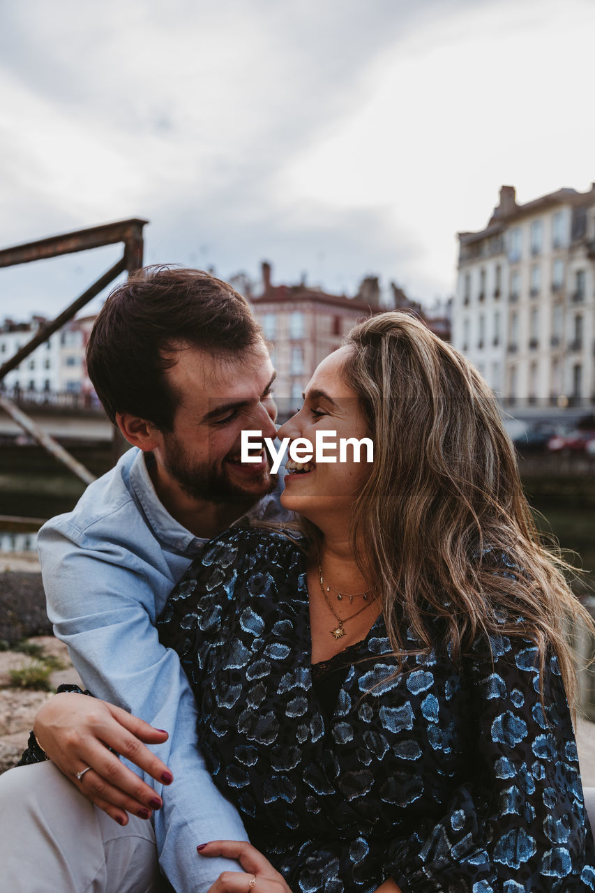 Positive young couple in casual clothes enjoying romantic date while sitting together on stone border in city with old buildings in background