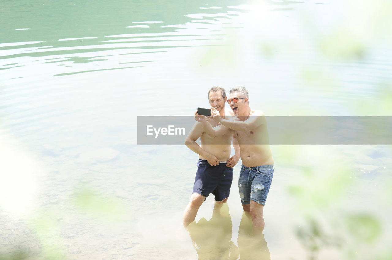 Shirtless gay couple taking selfie while standing in lake