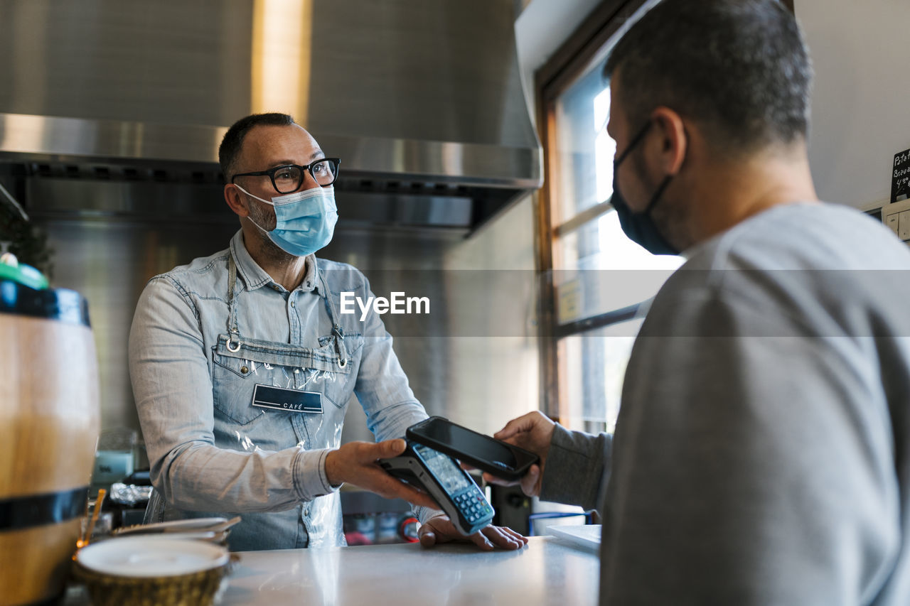 Waiter looking at male customer paying through smart phone at a bar