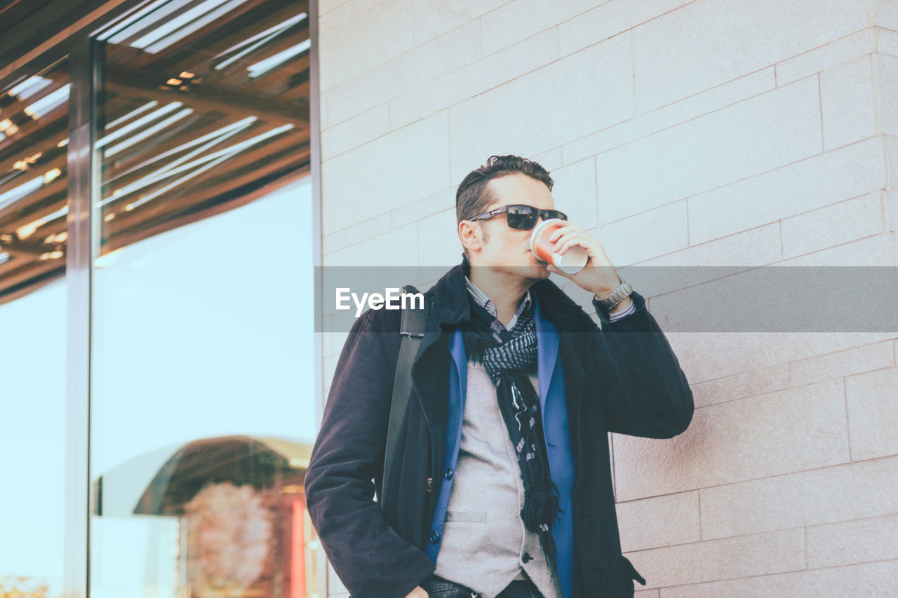 Fashionable man drinking coffee against wall outdoors