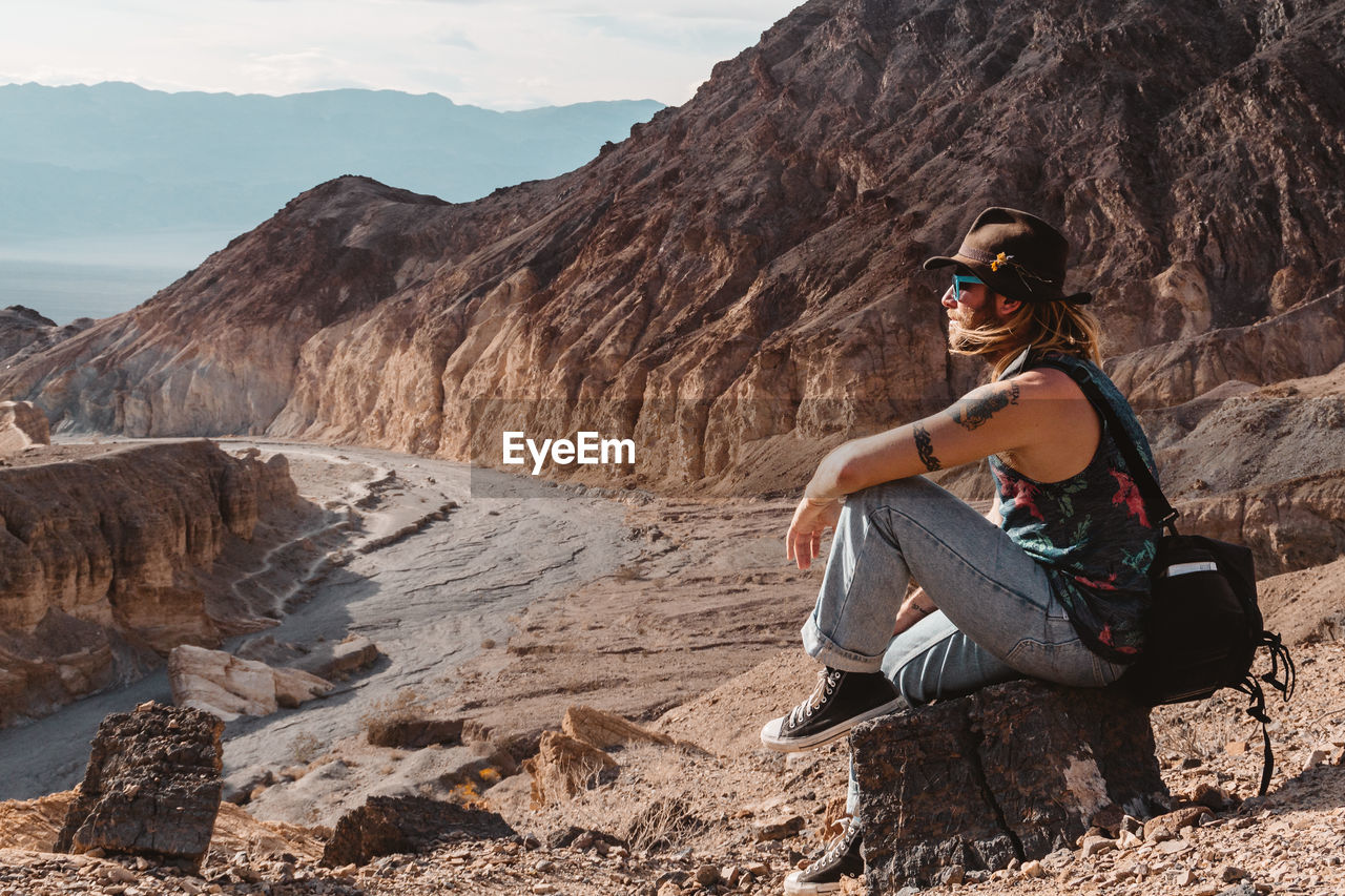 Male with hat and camera bag sitting on rocks in remote valley looking outward in death valley.