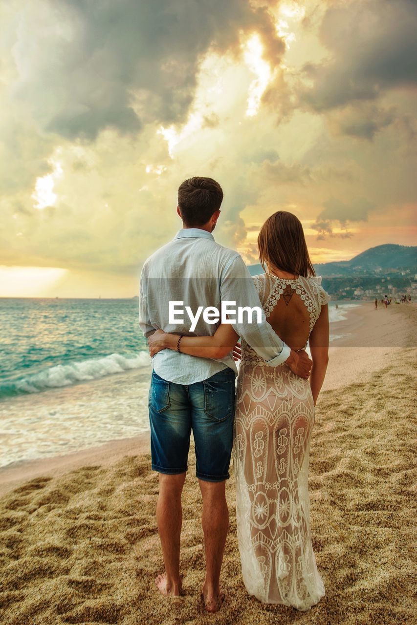 REAR VIEW OF FRIENDS STANDING ON BEACH DURING SUNSET