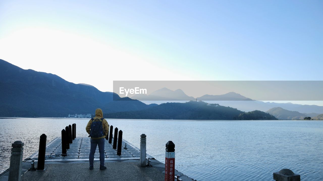 Rear view of man standing on pier over lake against clear sky