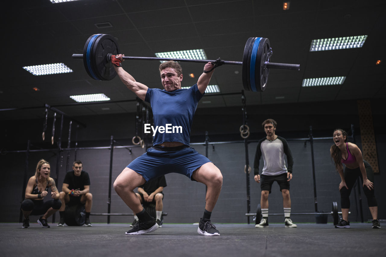 Athletes cheering man picking barbell while standing at gym
