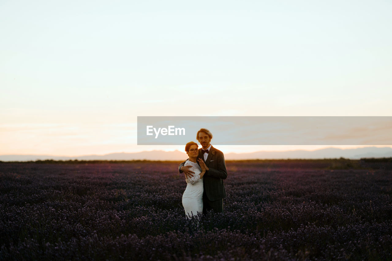 Side view of romantic newlywed couple standing face to face on spacious field against purple sunset sky looking at camera