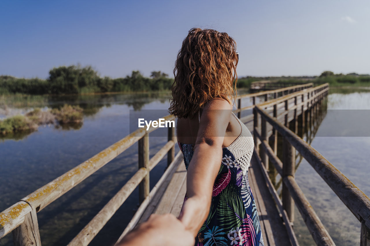 Woman holding hand of boyfriend while standing on footbridge against sky