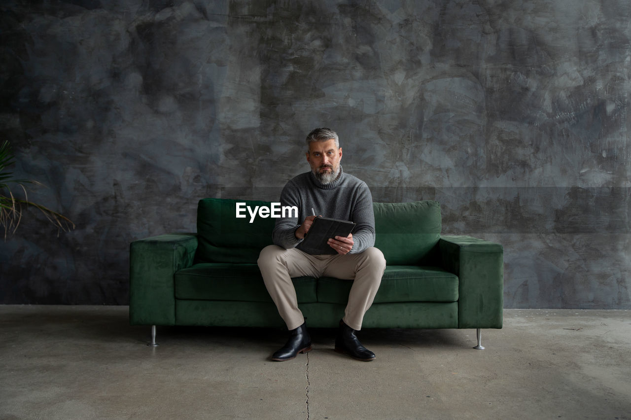 Portrait of handsome senior man sitting on green couch, using smartphone in stylish loft apartment