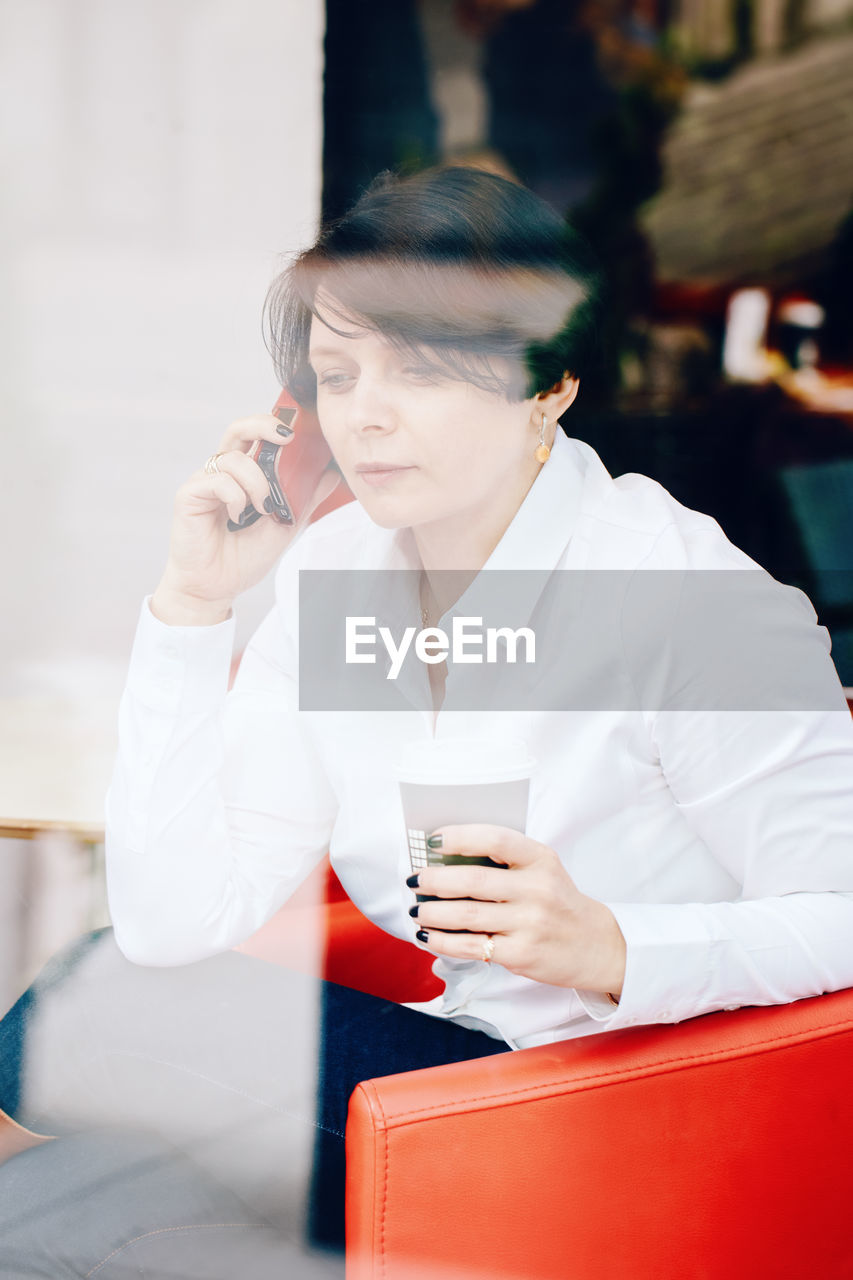 Woman talking over smart phone in cafe seen through window