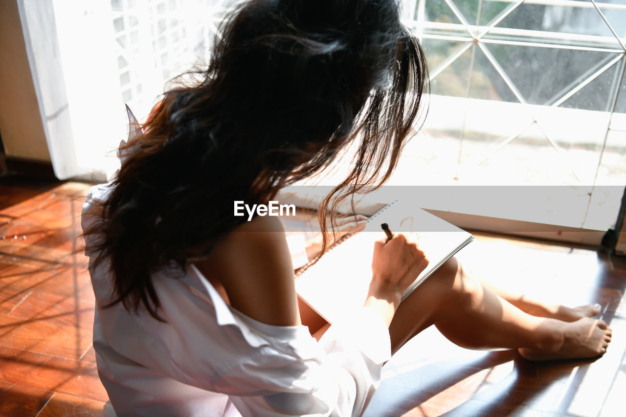 Rear view of woman writing on note pad while sitting on floor at home