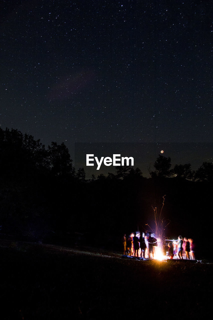 People around campfire on field against sky at night