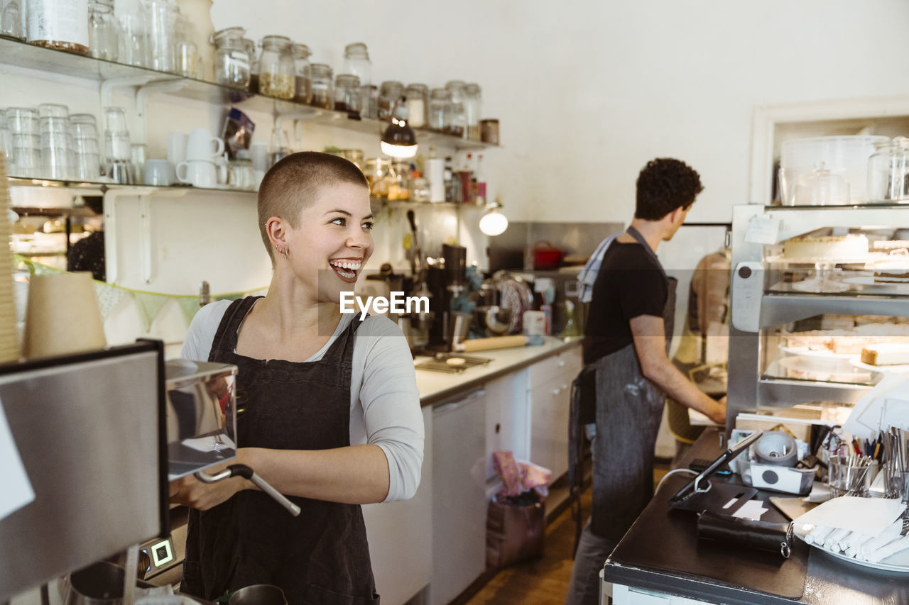 Young happy female owner looking away while preparing coffee in cafe