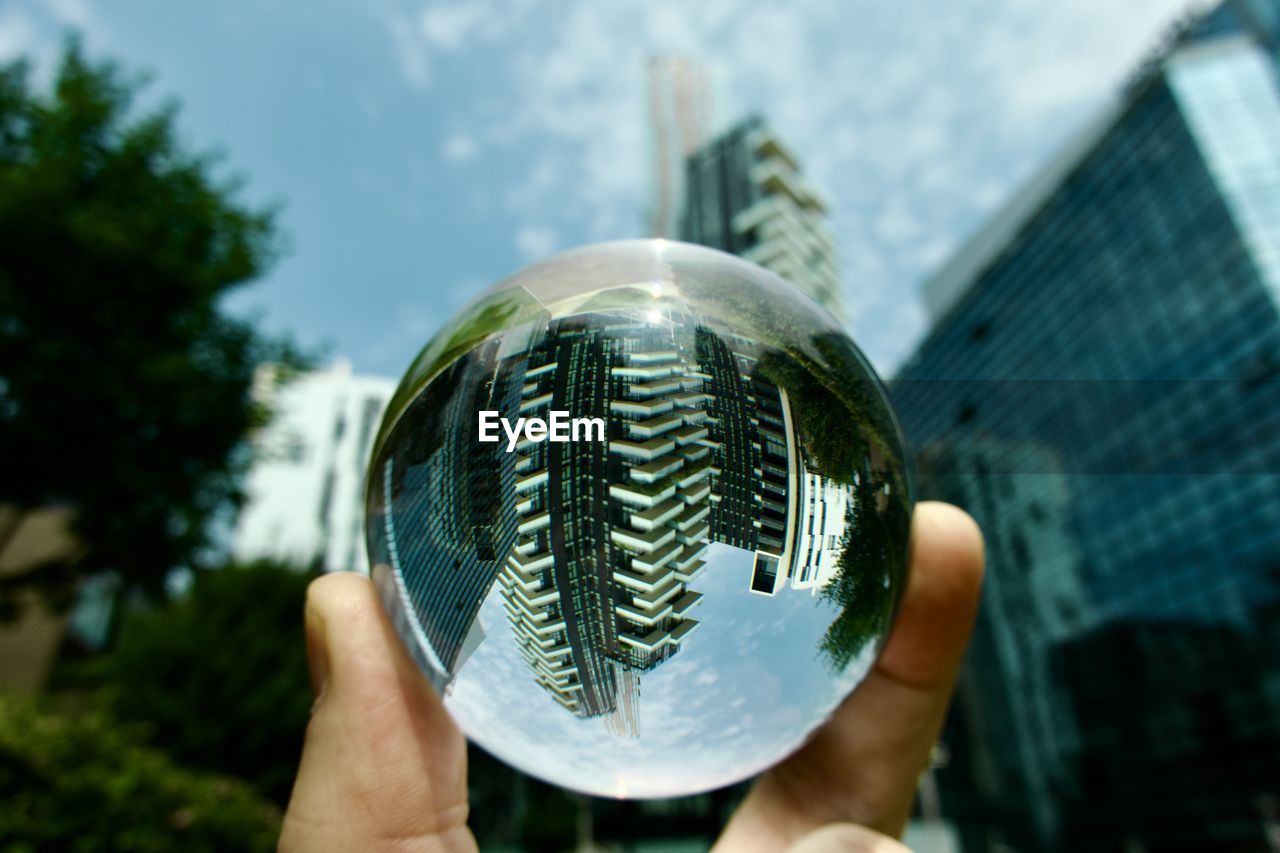 holding, hand, one person, architecture, building exterior, city, built structure, sphere, green, focus on foreground, nature, sky, day, close-up, reflection, outdoors, building, personal perspective, office building exterior, glass, adult, skyscraper, blue, tree, plant