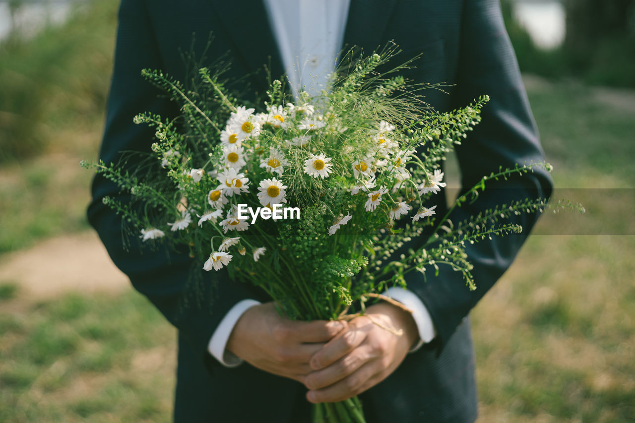 Midsection of bridegroom holding flowers while standing outdoors