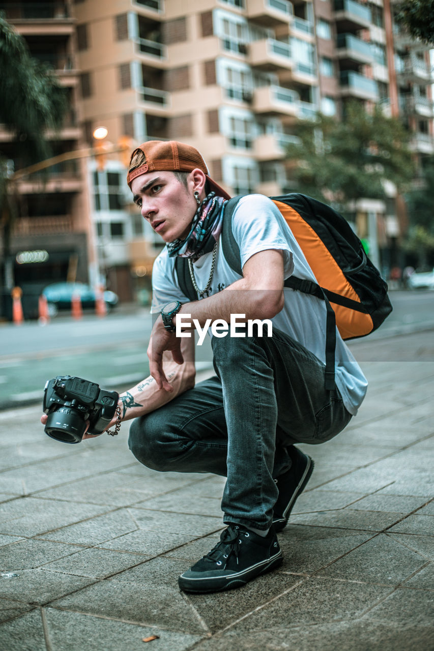 Young man looking away while holding camera on footpath in city