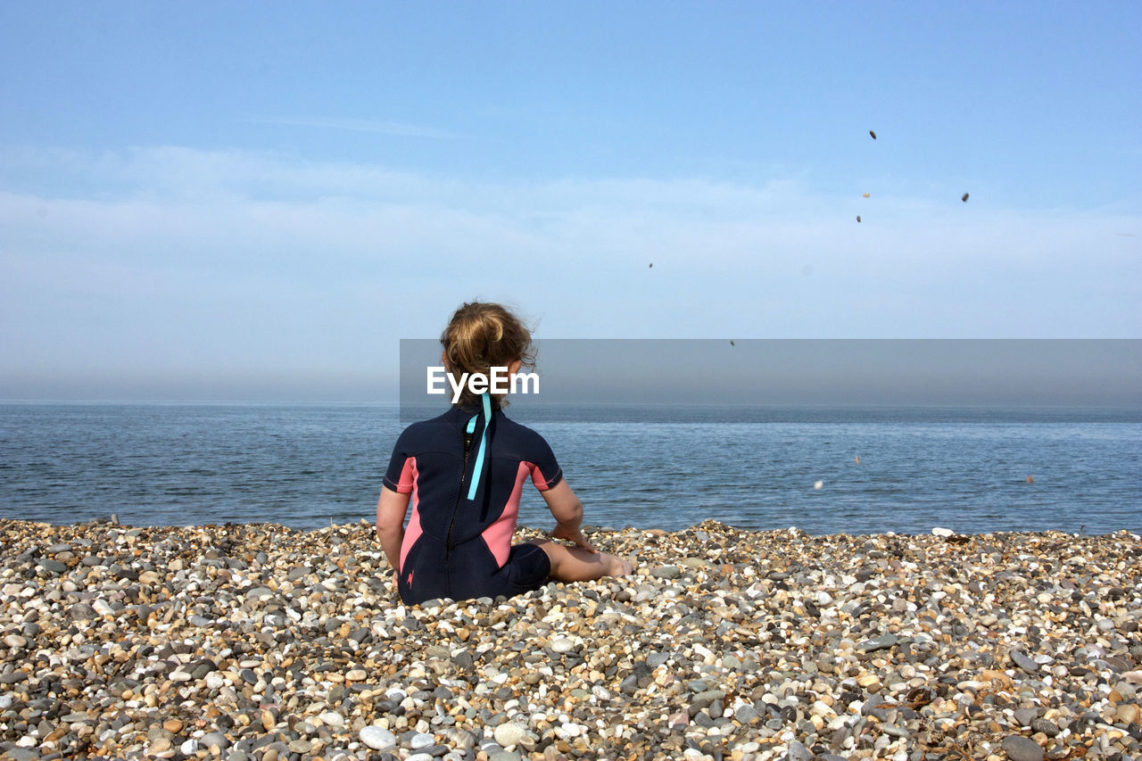 Girl sitting on pebbles at beach against sky