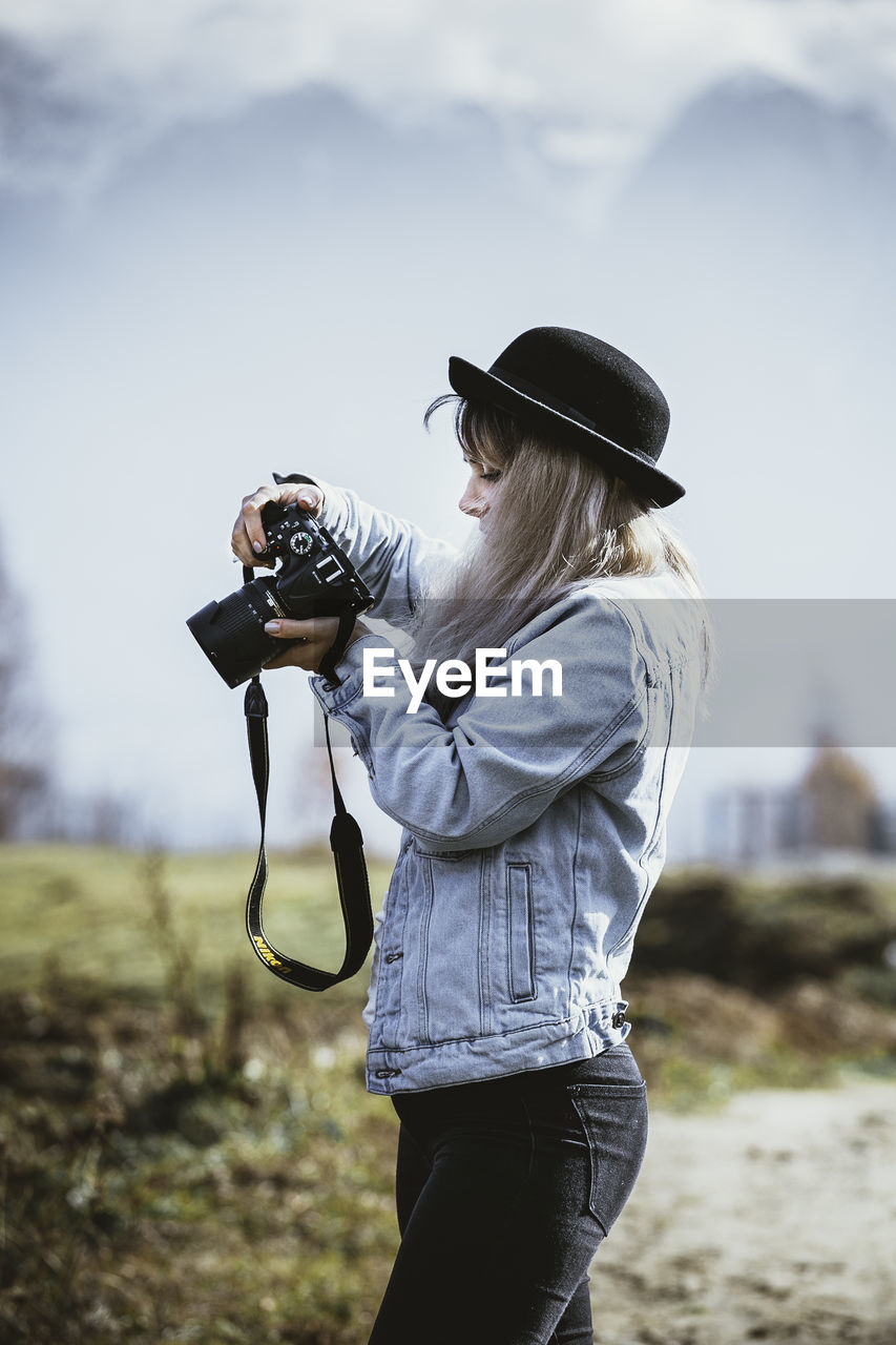 Side view of young woman photographing through camera against sky