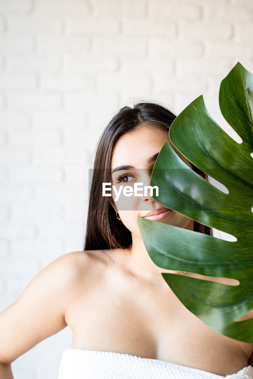 Beautiful brunette woman wearing bath towels holding a green monstera leaf in front of her face