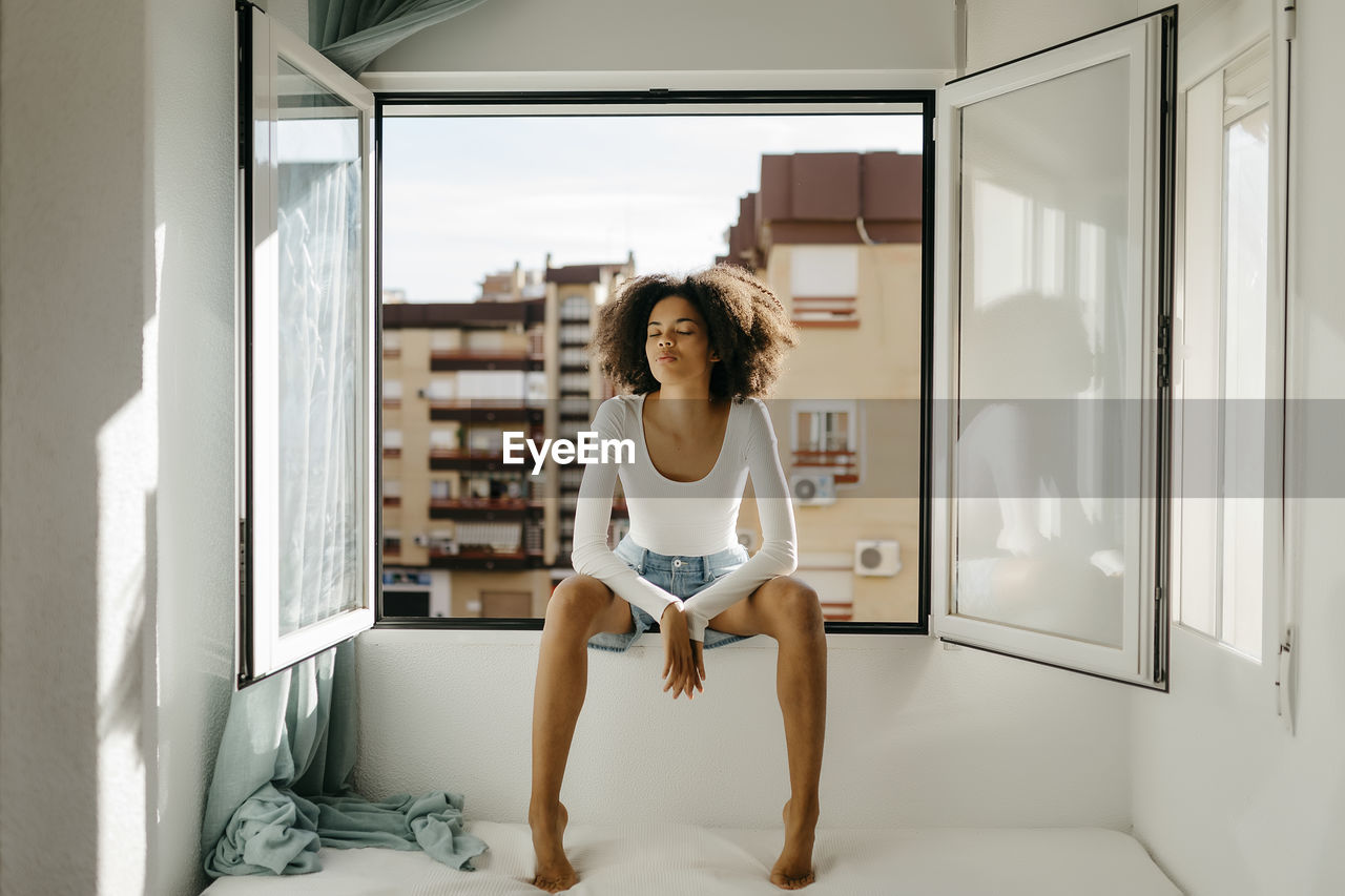 Mixed race woman with eyes closed sitting on window sill at home