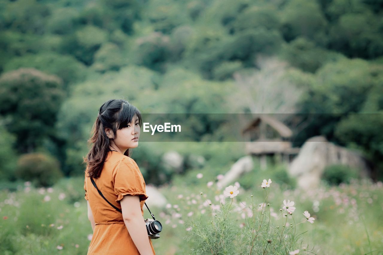 Beautiful young woman carrying camera while standing amidst plants on field