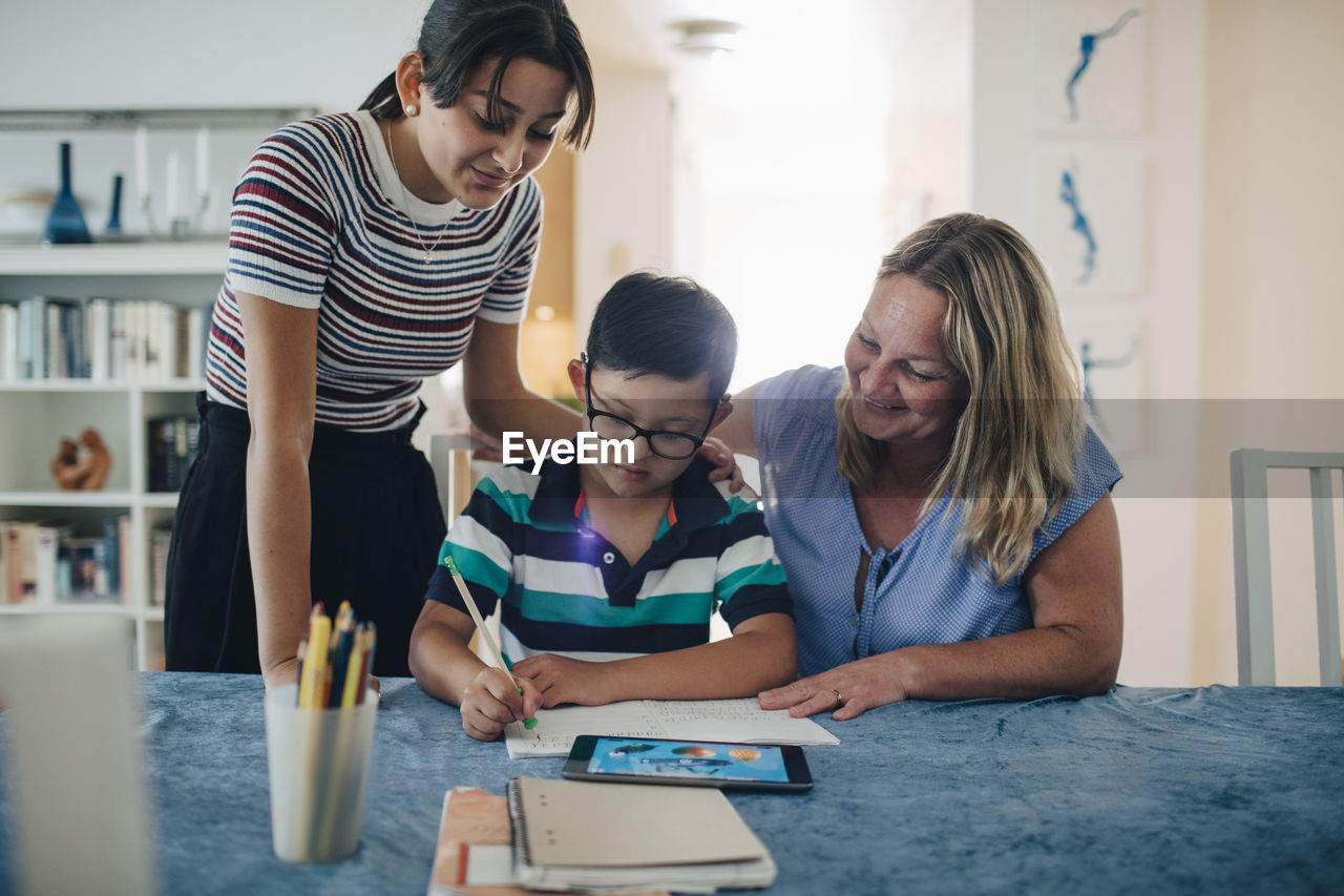 Smiling mother with teenage girl looking at boy studying at table