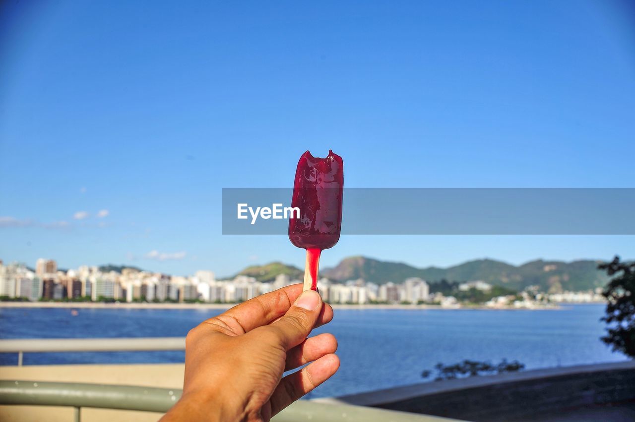 Cropped image of hand holding popsicle by lake against sky