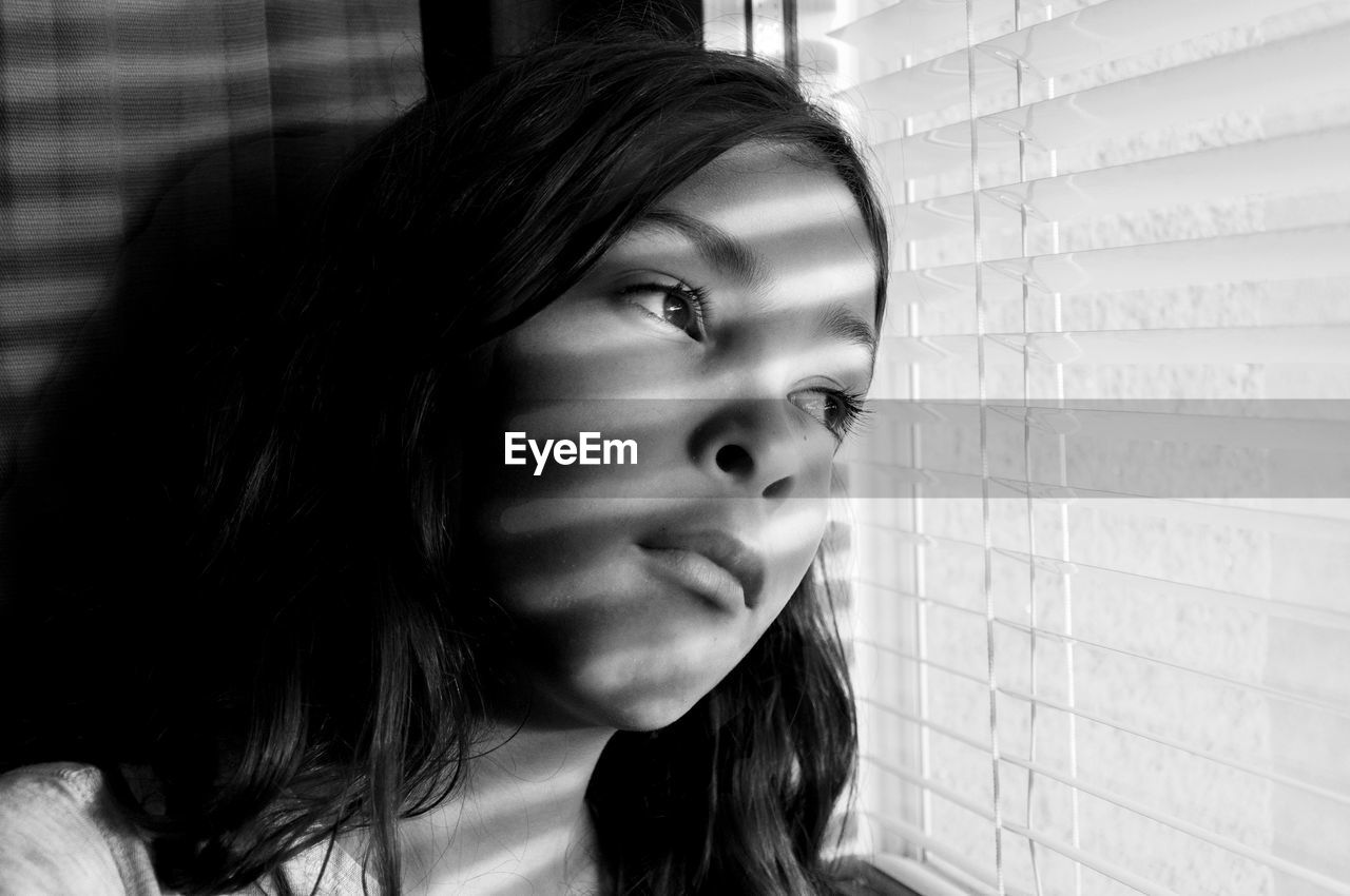 Close-up of lonely girl looking through window blinds at home