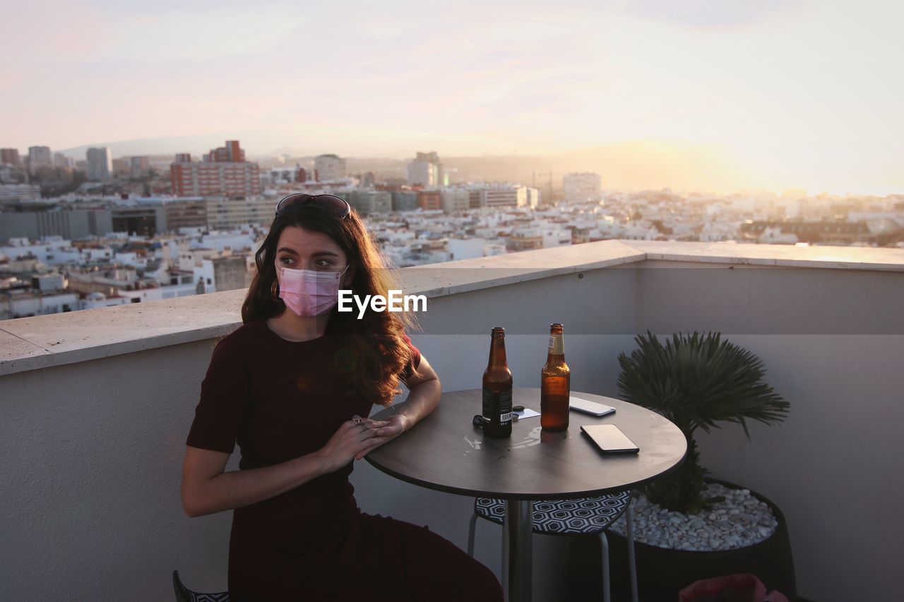 Portrait of young woman wearing a mask sitting against cityscape at sunset