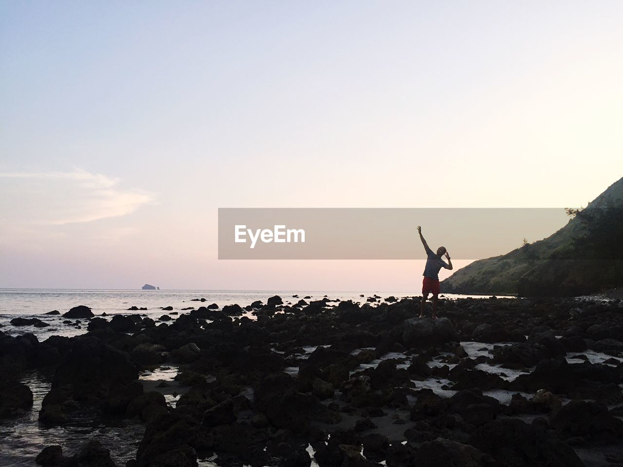 Mid distance view of man standing on rocky seashore against sky during sunset