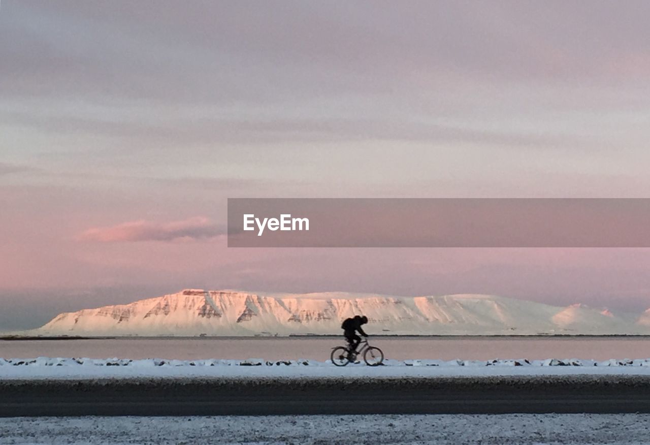 Man riding bicycle on snow field against sky during sunset