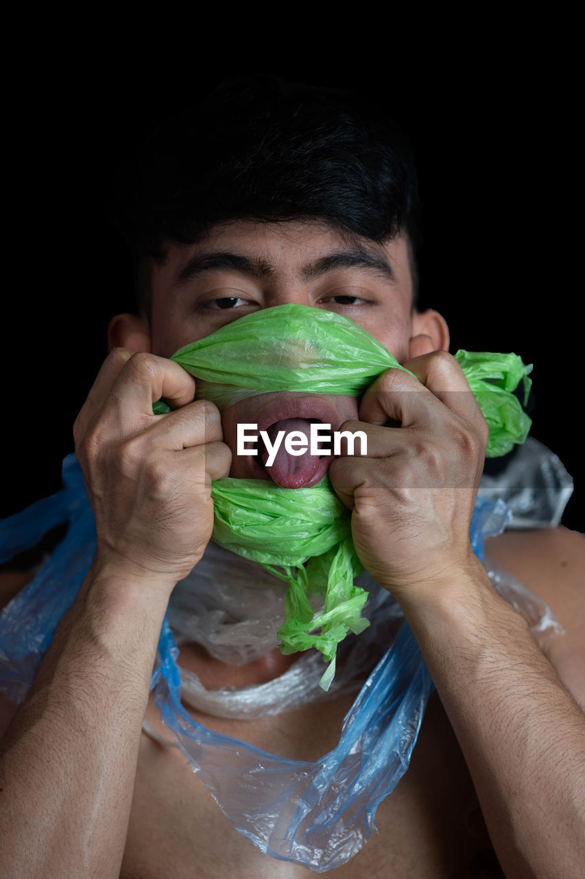 Portrait of shirtless young man with plastic covering face against black background