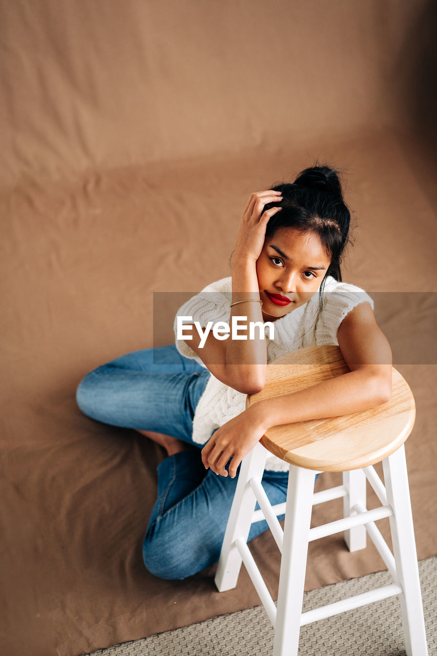 Full body of confident young hispanic lady wearing casual clothes sitting on the floor leaning on wooden stool in studio