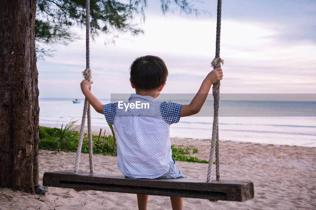 Rear view of boy swinging on rope swing at beach