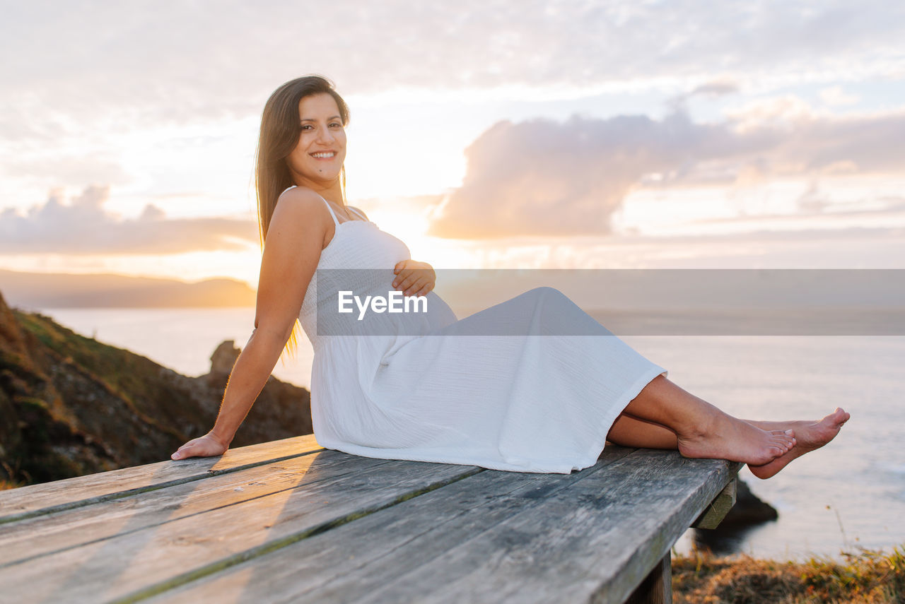 YOUNG WOMAN SITTING ON WOOD AGAINST SEA